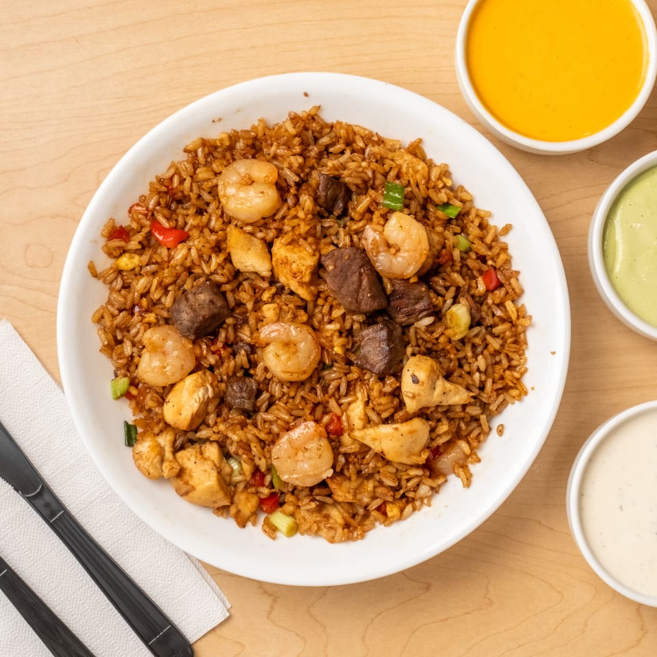 Overhead view of mixed fried rice with shrimp, chicken, beef, vegetables, dipping sauces, and black utensils on a wooden table.