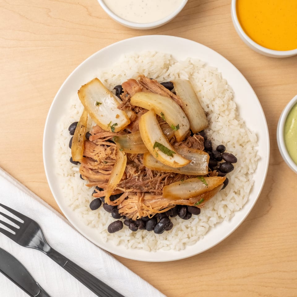 Plate of white rice topped with shredded pork, black beans, sautéed onions, and dipping sauces on a wooden table.