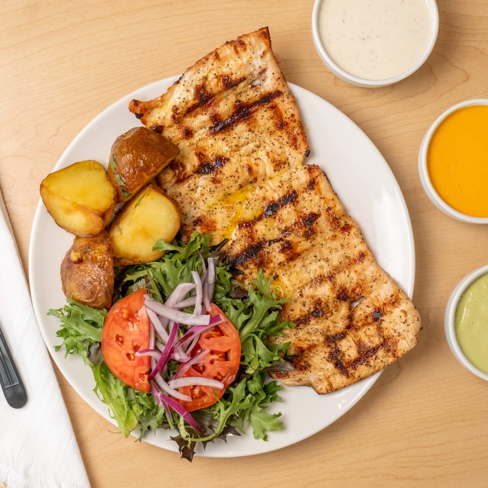 Grilled chicken platter with roasted potatoes, mixed green salad, and three dipping sauces on a wooden table.