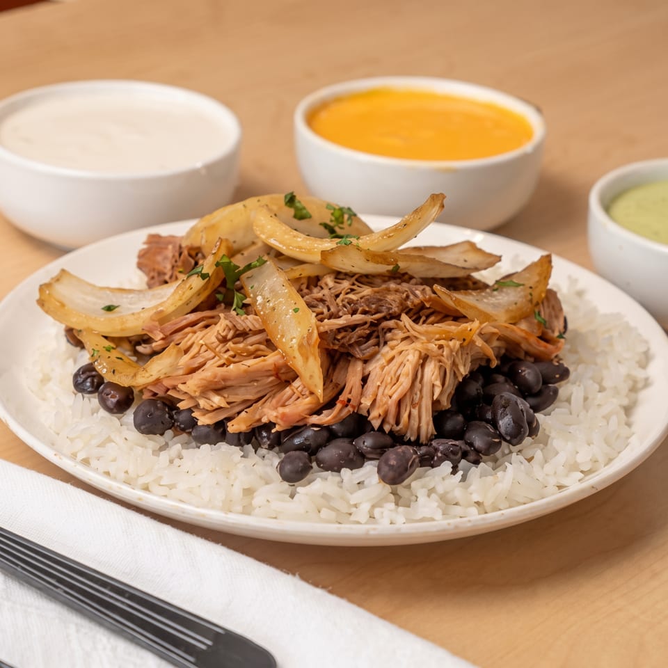 Plate of white rice, black beans, shredded meat, caramelized onions, and sauces on a wooden table.