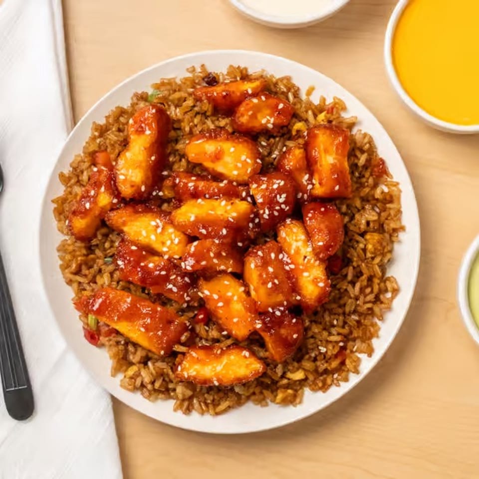 Plate of fried rice topped with glazed orange chicken pieces and sesame seeds, surrounded by dipping sauces and utensils.