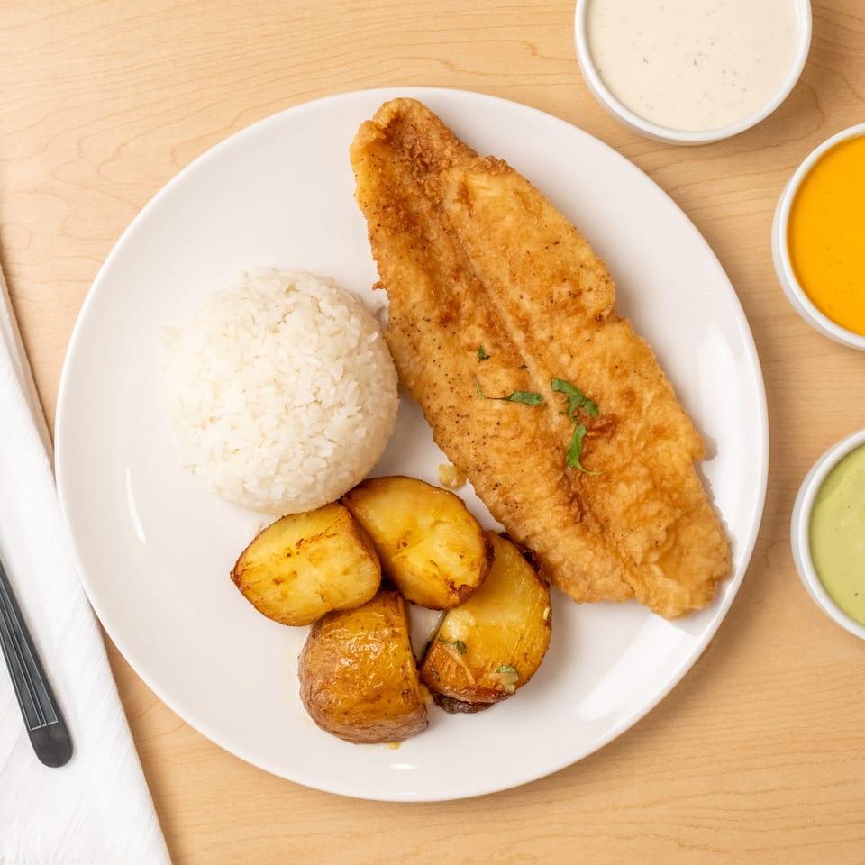 Plate of crispy fried fish, white rice, roasted potatoes, and three dipping sauces on a wooden table.