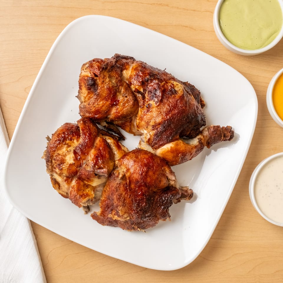 Roasted chicken pieces on a white platter with green, orange, and creamy dipping sauces on a wooden table.