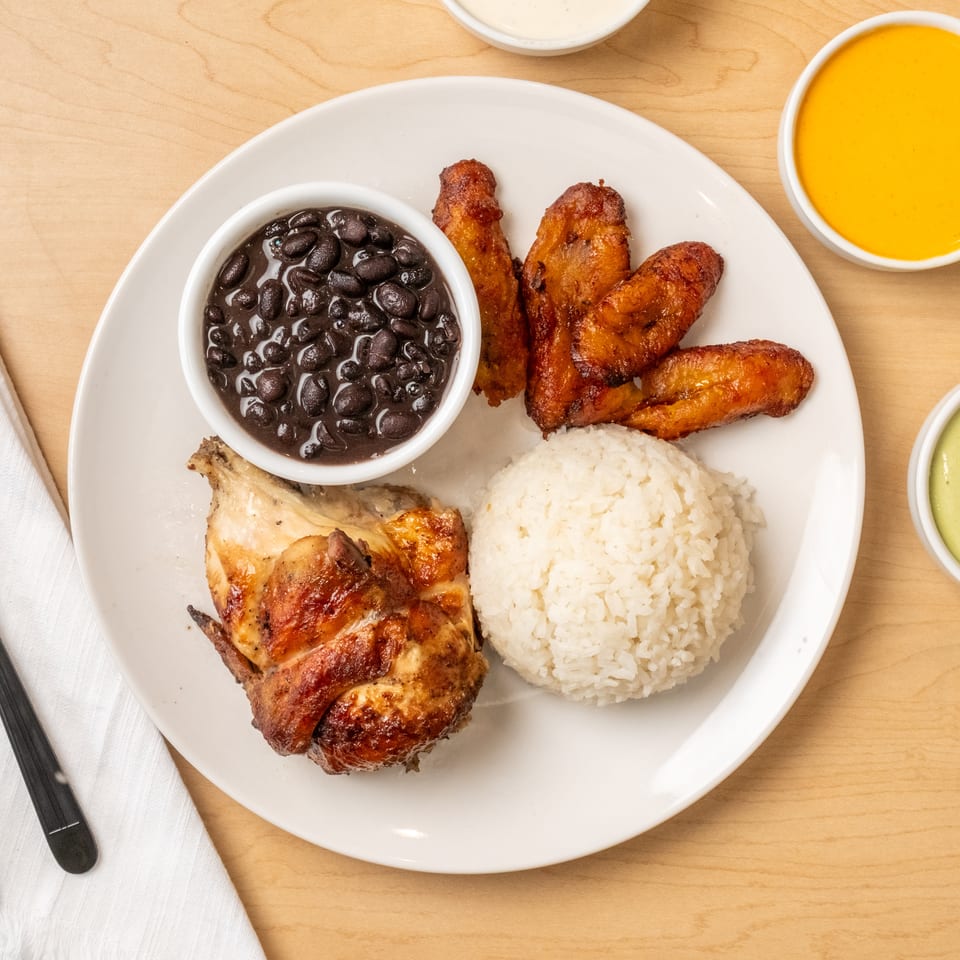 Plate of grilled chicken, white rice, black beans, fried plantains, and colorful sauces on a wooden table.