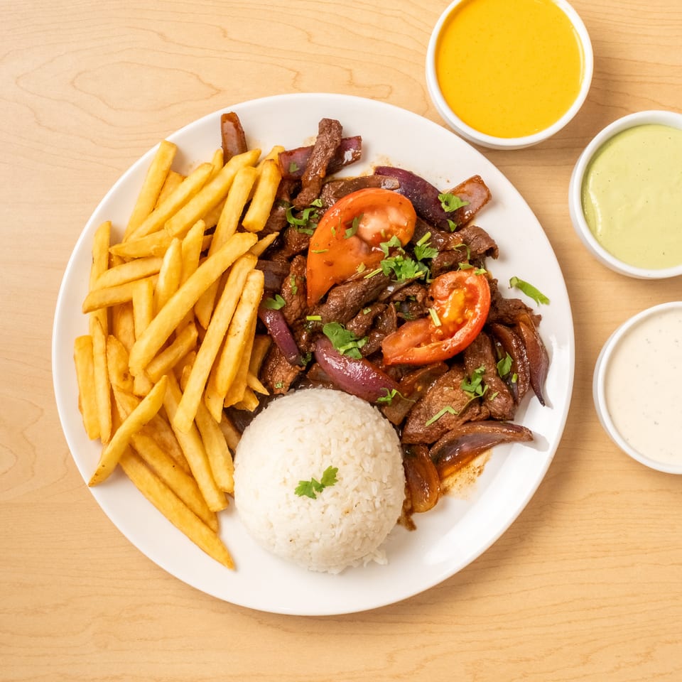 Plate of sautéed beef with onions and tomatoes, white rice, French fries, and three dipping sauces on a wooden table.