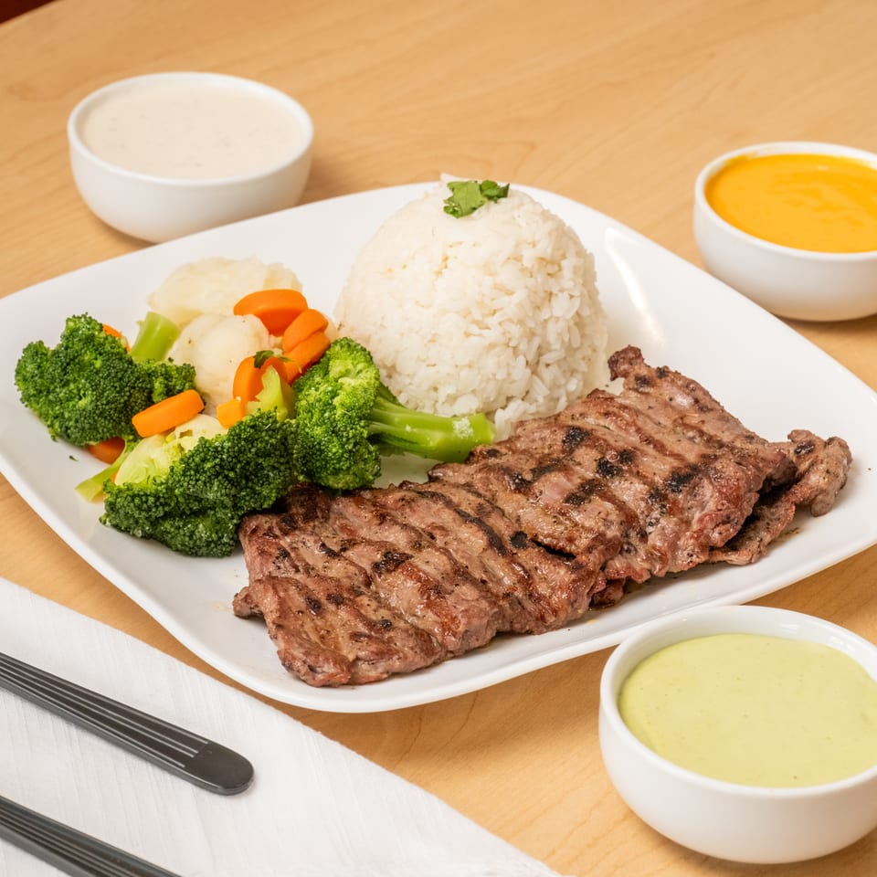 Grilled steak with white rice, steamed broccoli and carrots, accompanied by three dipping sauces on a white plate.